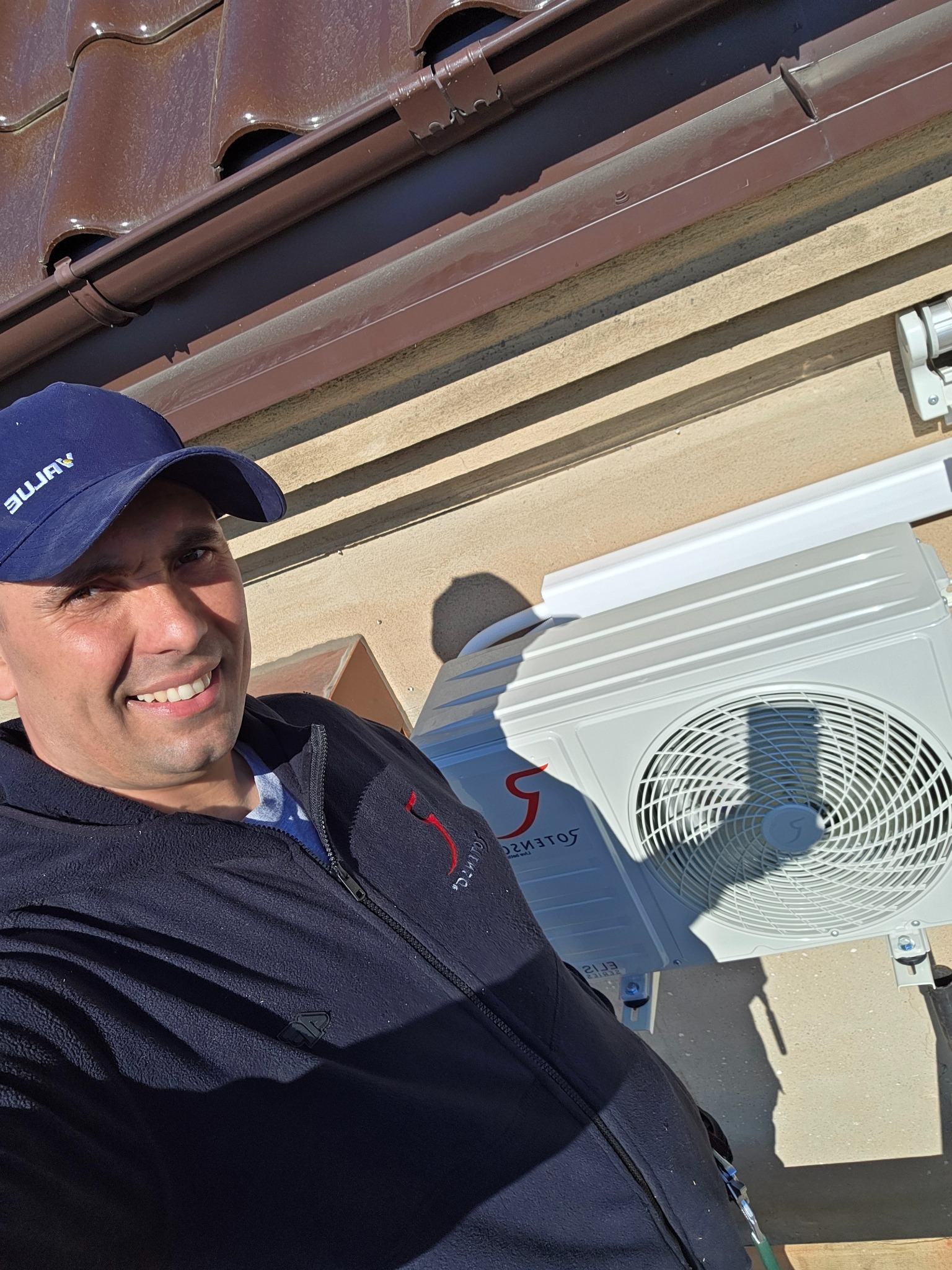 Uśmiechnięty mężczyzna w czapce pozuje obok zamontowanej jednostki zewnętrznej klimatyzacji na elewacji budynku z brązowym dachem. Widoczne mocowania i przewody.