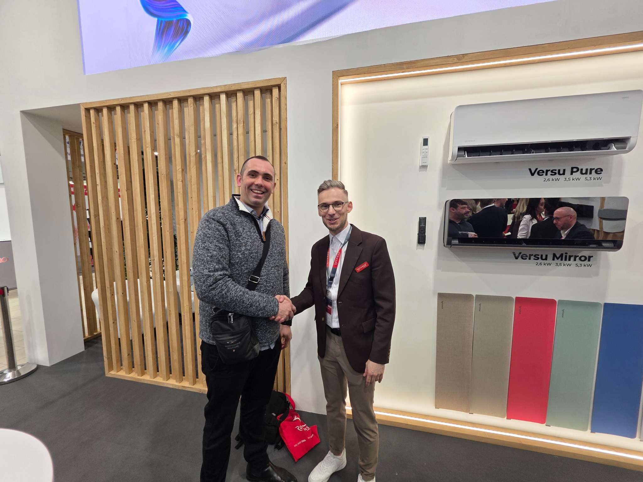 Dwóch mężczyzn wita się na tle ekspozycji klimatyzacji Versu Pure i Versu Mirror z panelami w różnych kolorach. Widoczny model klimatyzatora i parametry techniczne.