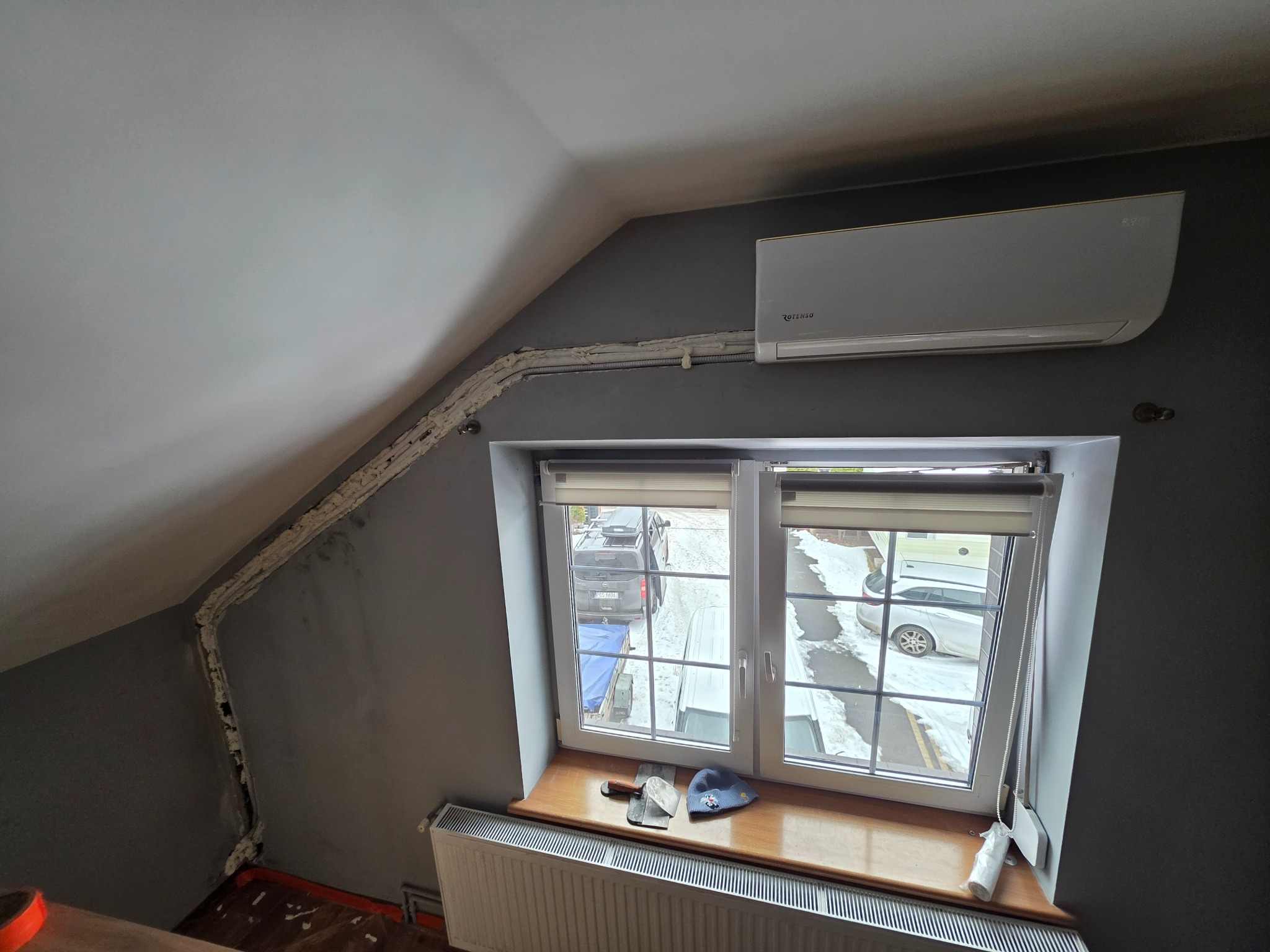Klimatyzator Rotenso zamontowany w pokoju na poddaszu, widoczne rury zabezpieczone pianką montażową, okno z widokiem na zewnątrz, narzędzia na parapecie.