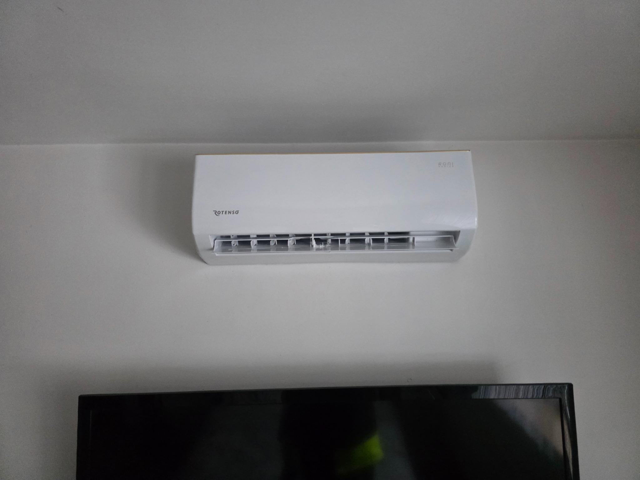 Biały klimatyzator Rotenso zamontowany na ścianie nad czarnym telewizorem. Widoczne detale obudowy i wlotu powietrza. Minimalistyczne wnętrze, neutralne oświetlenie.