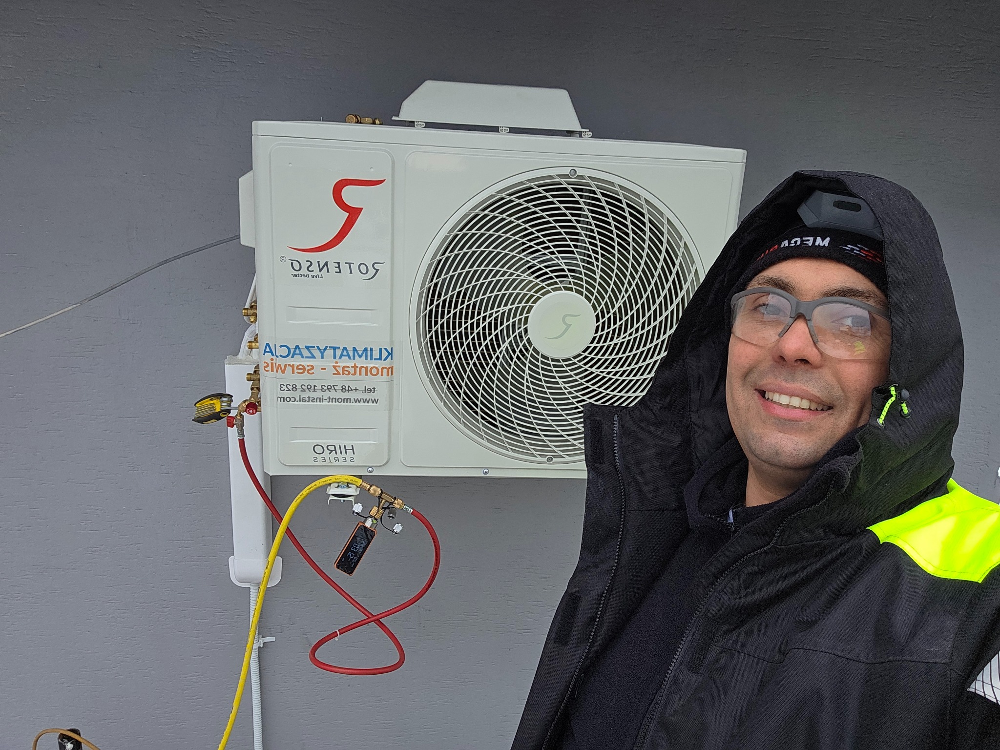Instalator uśmiecha się obok zamontowanej klimatyzacji Rotenso HIRO z podłączonymi przewodami serwisowymi i manometrem. Widoczny adres strony internetowej.