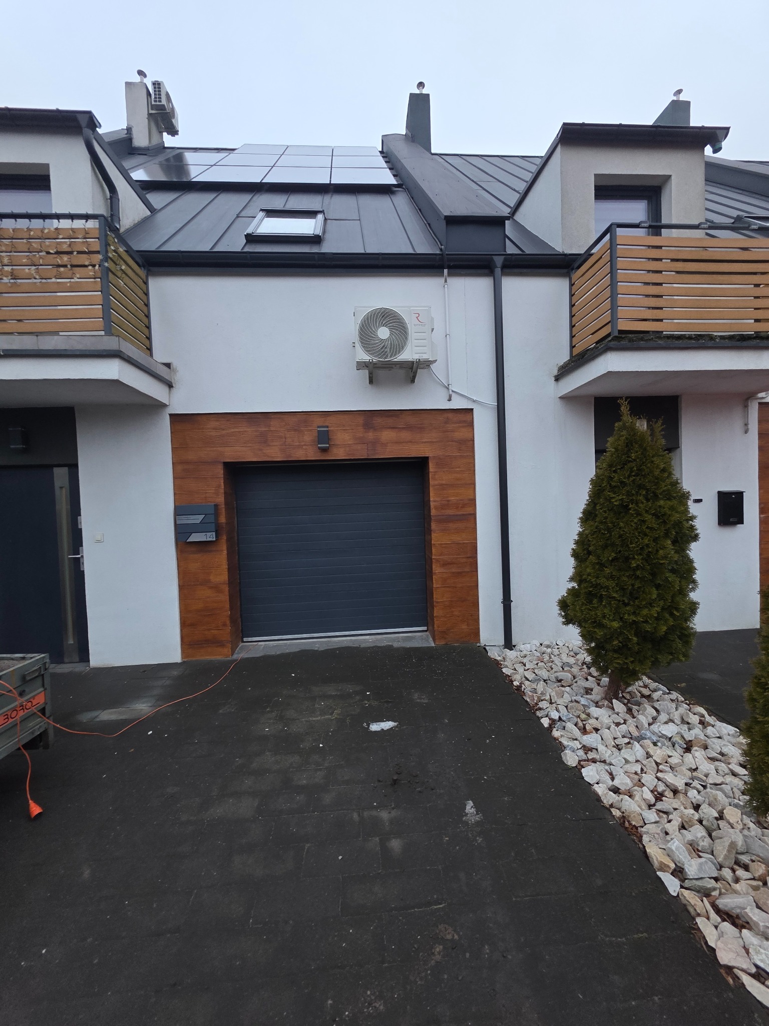 Biały dom z szarą bramą garażową i klimatyzatorem na elewacji. Na dachu panele słoneczne i okno dachowe. Widoczne elementy drewniane i ozdobne kamienie.