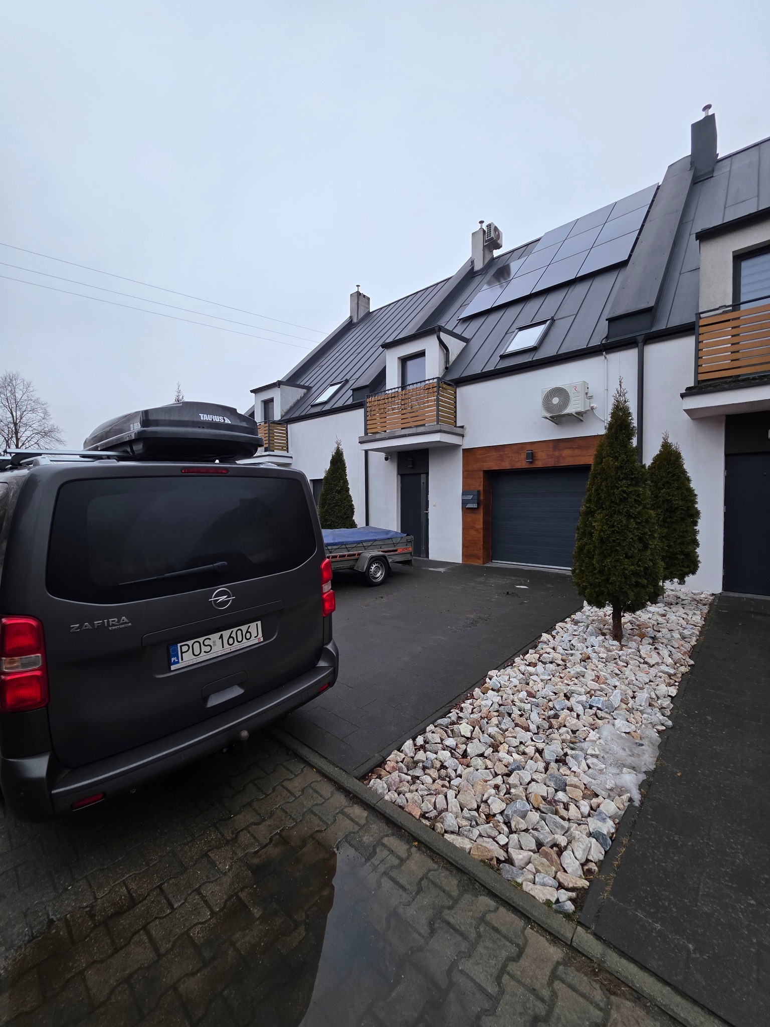 Nowoczesny dom szeregowy z klimatyzacją na elewacji, panele słoneczne na dachu. Na podjeździe zaparkowany samochód z bagażnikiem dachowym.