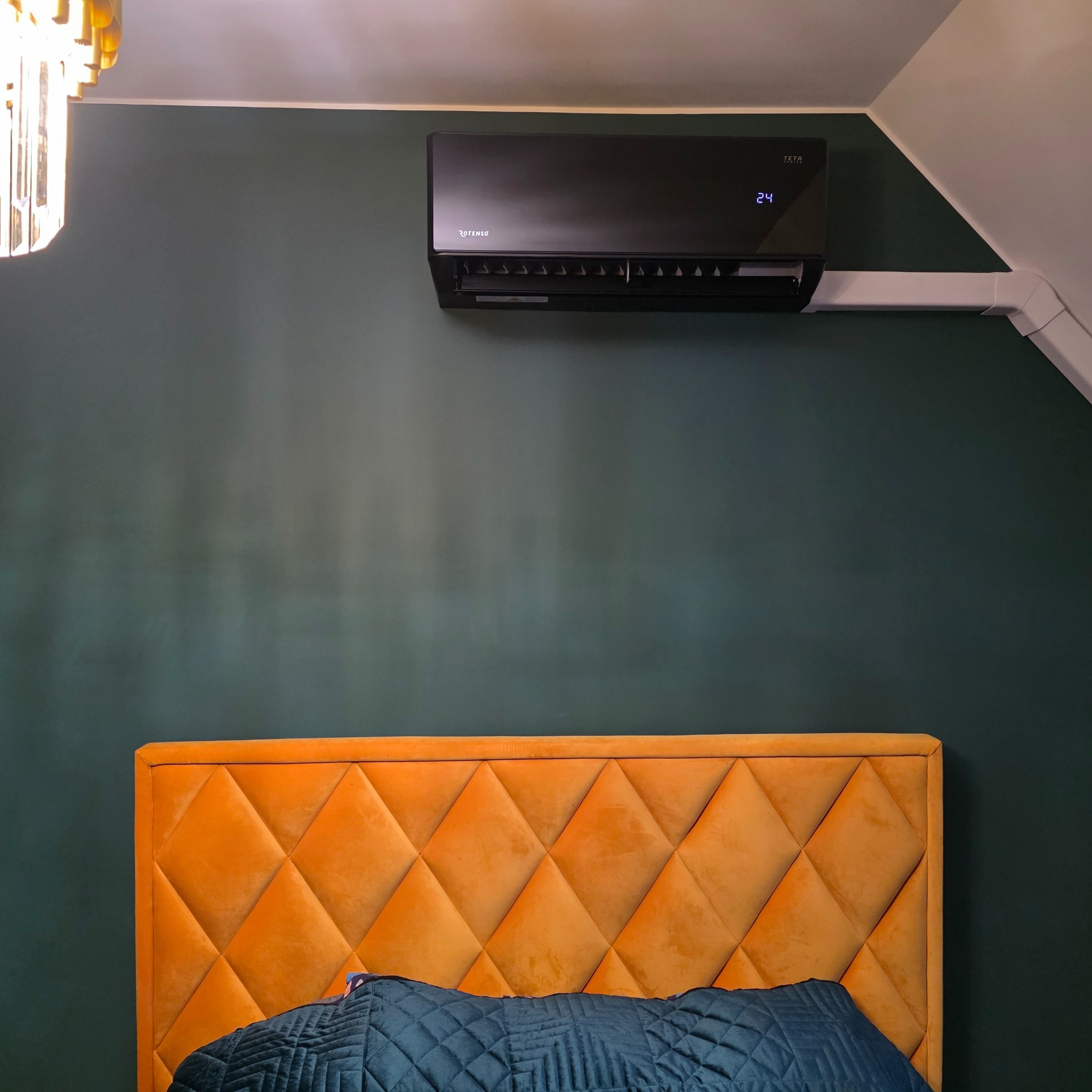 Czarny klimatyzator Rotenso zamontowany na ciemnozielonej ścianie nad pomarańczowym zagłówkiem łóżka. Widoczne białe rury instalacyjne i fragment sufitu.