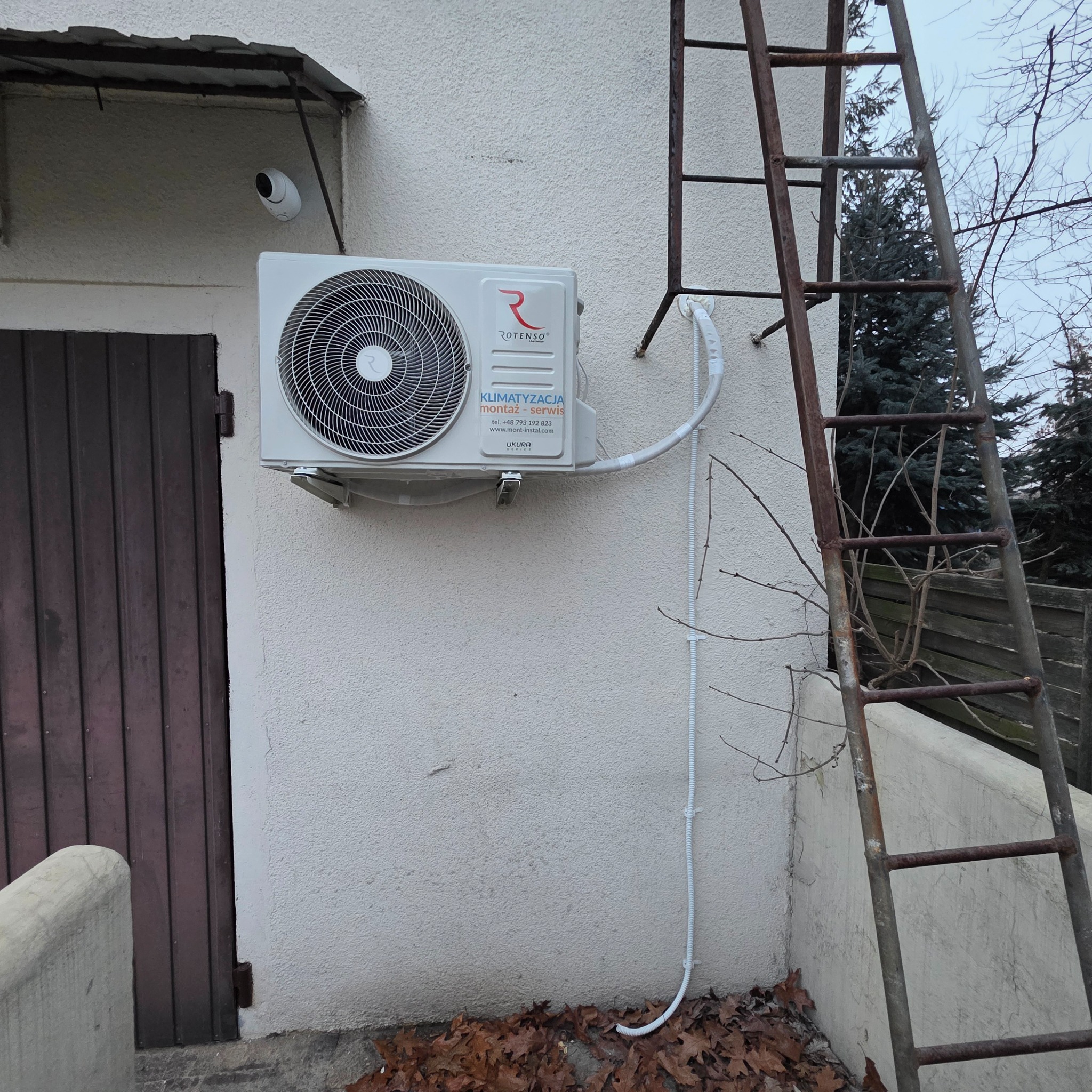Zamontowany klimatyzator Rotenso na elewacji budynku, z widocznymi przewodami i starą, metalową drabiną opartą o ścianę. Widoczne liście na ziemi.