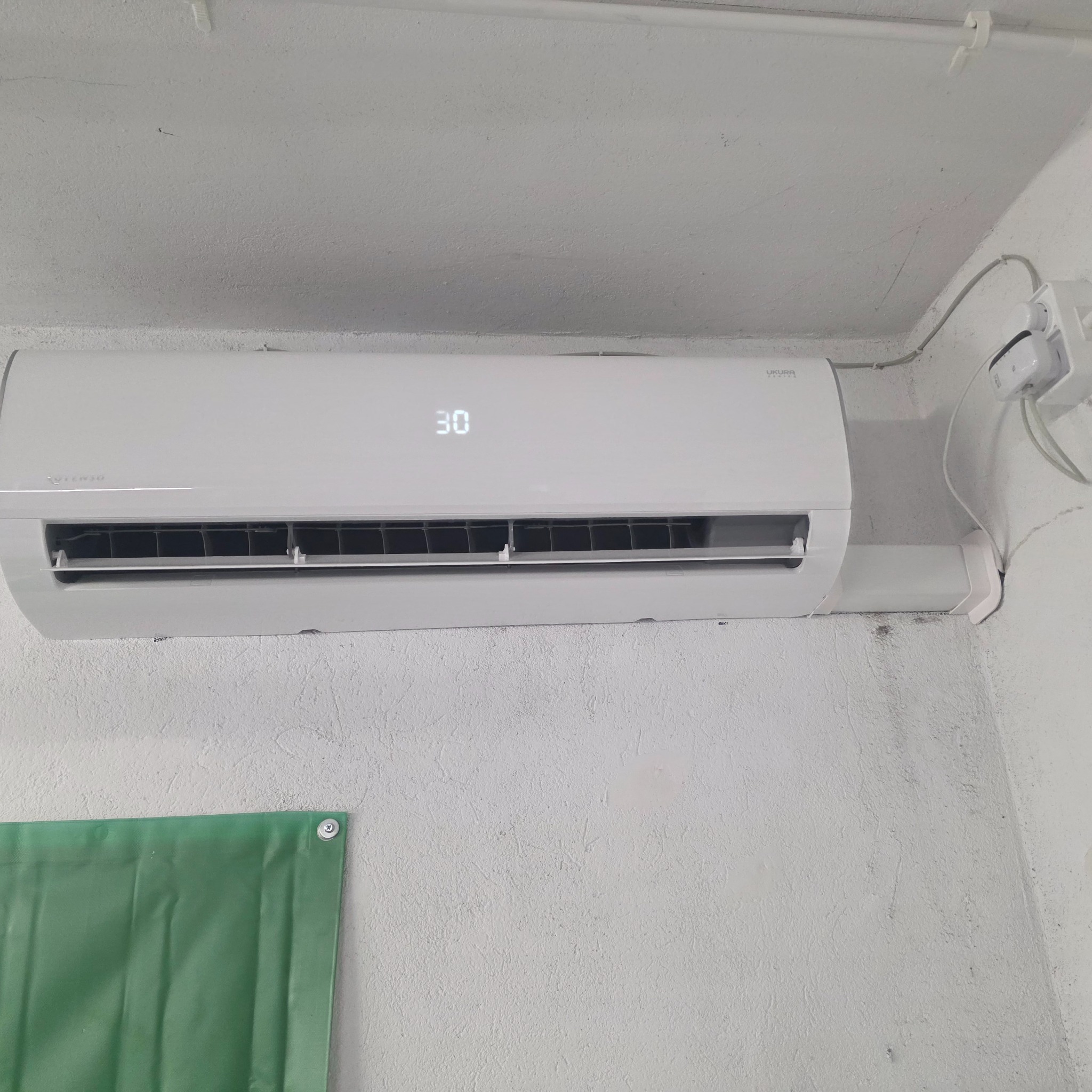 Biały klimatyzator Ukura z wyświetlaczem 30 stopni Celsjusza, zamontowany na białej, teksturowanej ścianie. Widoczne przewody i gniazdko elektryczne.