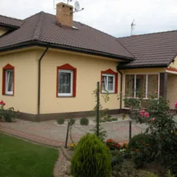 Żółty dom jednorodzinny z brązowym dachem i czerwonymi obramowaniami okien, otoczony zadbanym ogrodem z różami i ścieżką z kostki brukowej.