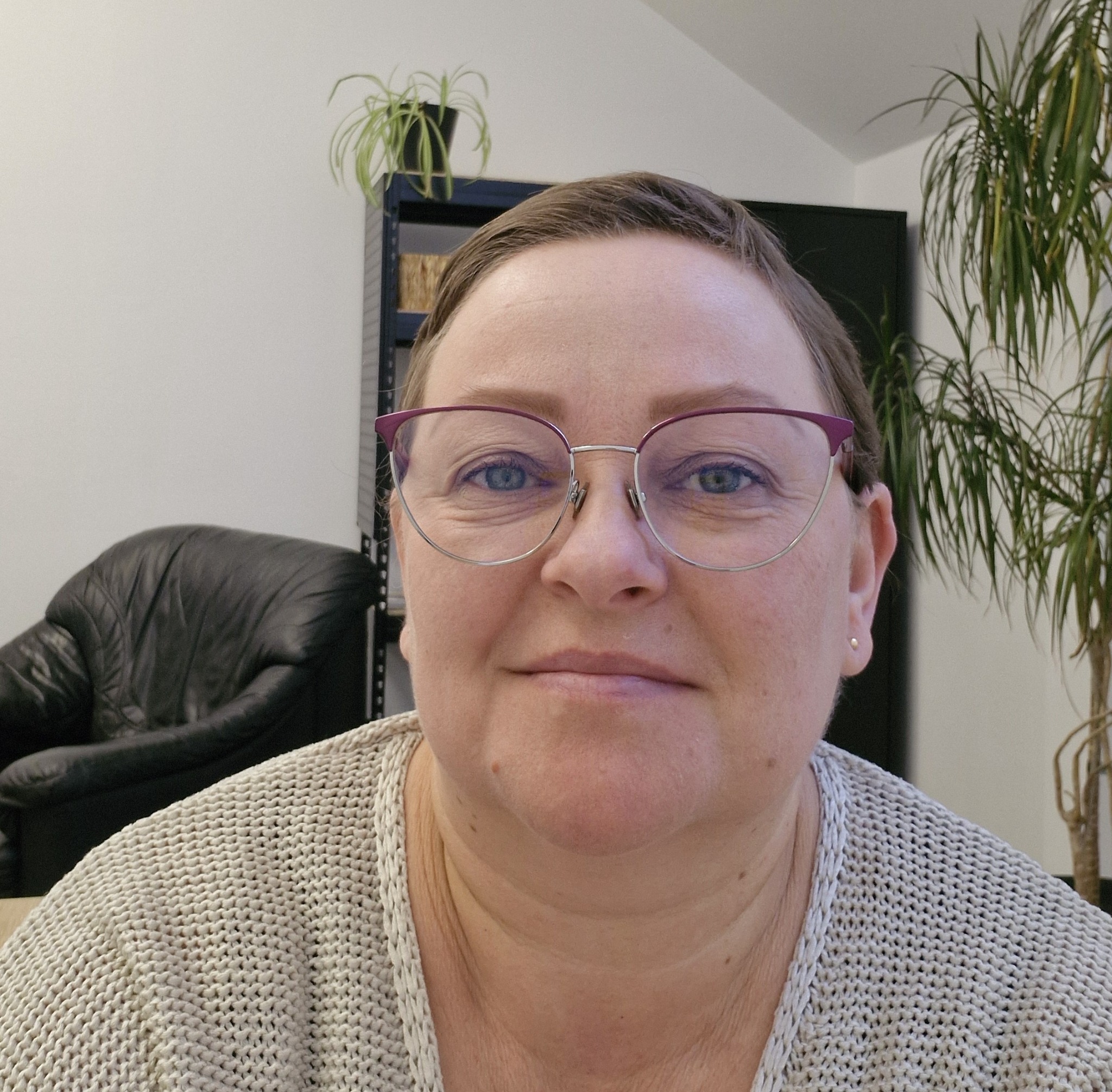 Portret kobiety w okularach z fioletowymi oprawkami, w biurowym otoczeniu z roślinami i czarnym fotelem. Uśmiechnięta twarz, naturalne światło.