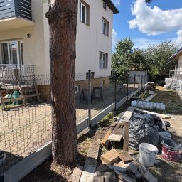 Kamil Grabowiec - Częściowo zainstalowane ogrodzenie panelowe z betonową podmurówką przed budynkiem mieszkalnym, z dużym drzewem na pierwszym planie i materiałami budowlanymi na ziemi.