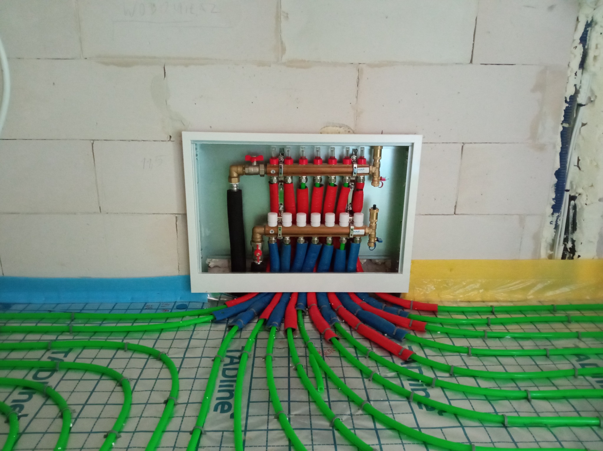 Rozdzielacz ogrzewania podłogowego z rurami w kolorze zielonym, czerwonym i niebieskim, zamontowany na ścianie z białych bloczków, widok z góry.