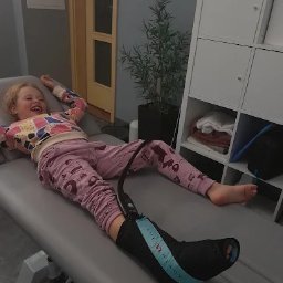 Tu. Fizjoterapia Tuszyn - fizjoterapia - terapia manualna - trening medyczny - Uśmiechnięta dziewczynka leży na leżance z ortezą na nodze. Widoczny fragment pomieszczenia z szafką i rośliną doniczkową.