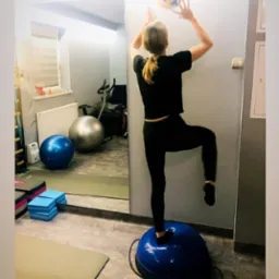 Kobieta ćwiczy równowagę na bosu, podrzucając piłkę. W tle lustro z widokiem na sprzęt do ćwiczeń. Trening personalny w studio.