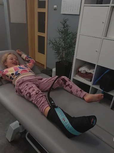 Uśmiechnięta dziewczynka leży na leżance z ortezą na nodze. Widoczny fragment pomieszczenia z szafką i rośliną doniczkową.