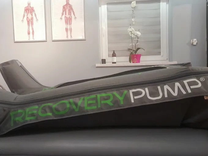 Aparat do presoterapii Recovery Pump na stole w gabinecie. Na ścianie plakaty anatomiczne i kwiat. Sprzęt do masażu limfatycznego w gabinecie fizjoterapii.