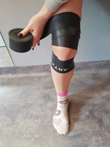 Owijanie kolana elastyczną, czarną taśmą. Widoczna noga w skarpetce z motywem pączków. Fragment dłoni trzymającej zwiniętą taśmę. Szary mur w tle.