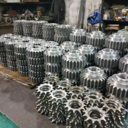 Viktor Komissarov - Stosy obrobionych, metalowych kół zębatych na warsztatowym stole. Widoczne narzędzia w tle. Szare, metaliczne elementy ułożone w rzędach.