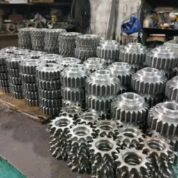 Stosy obrobionych, metalowych kół zębatych na warsztatowym stole. Widoczne narzędzia w tle. Szare, metaliczne elementy ułożone w rzędach.