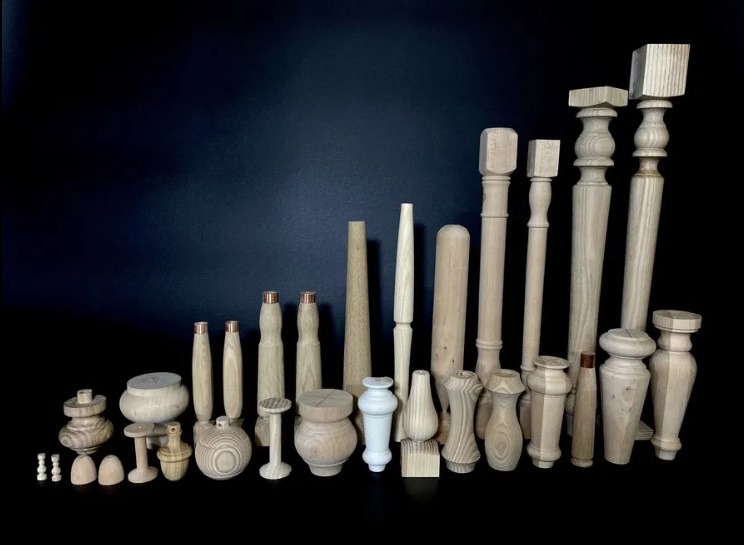 Różnorodne, surowe elementy toczone z jasnego drewna, niektóre z miedzianymi akcentami, ułożone na ciemnym tle. Widoczne detale rzemiosła.