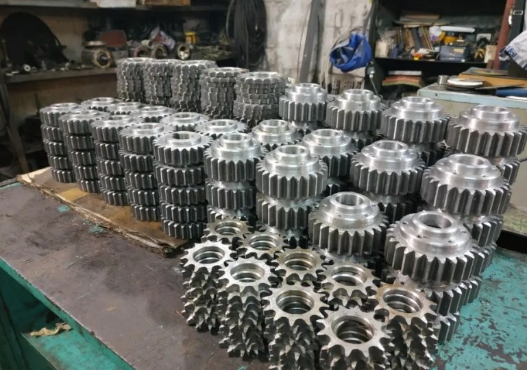 Stosy obrobionych, metalowych kół zębatych na warsztatowym stole. Widoczne narzędzia w tle. Szare, metaliczne elementy ułożone w rzędach.