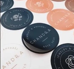 Zbliżenie na okrągłe naklejki z minimalistycznym logo i napisem 'Chandra Jewelry & Apparel ESTD. 2015' w kolorach granatu, różu i bieli. Widoczne refleksy światła.