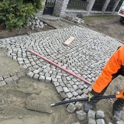 Aleks Kołek - Układanie kostki granitowej 10 cm w łuk. Widoczny fragment ułożonej nawierzchni, poziomica, narzędzia i bryły kostki. Praca w toku, pomarańczowa kurtka.