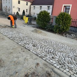 Aleks Kołek - Układanie kostki granitowej na posesji. Widoczne ułożone fragmenty i pracownik w pomarańczowej kurtce. W tle domy i roślinność.