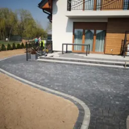 Ułożona kostka brukowa w kolorze grafitowym z jasną obwódką, prowadząca do domu z balkonem. Widoczny fragment trawnika i osoby w tle.