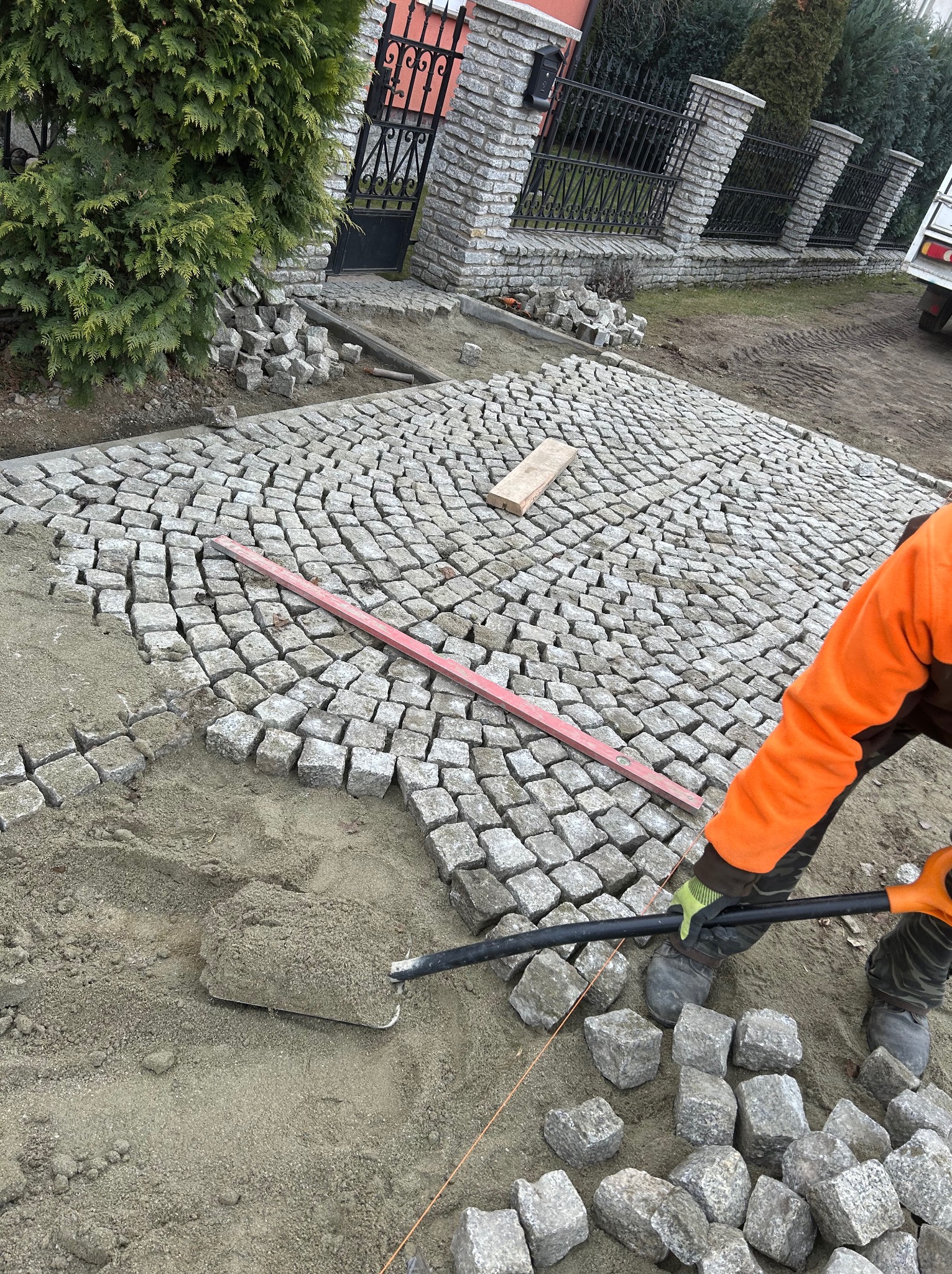 Układanie kostki granitowej 10 cm w łuk. Widoczny fragment ułożonej nawierzchni, poziomica, narzędzia i bryły kostki. Praca w toku, pomarańczowa kurtka.