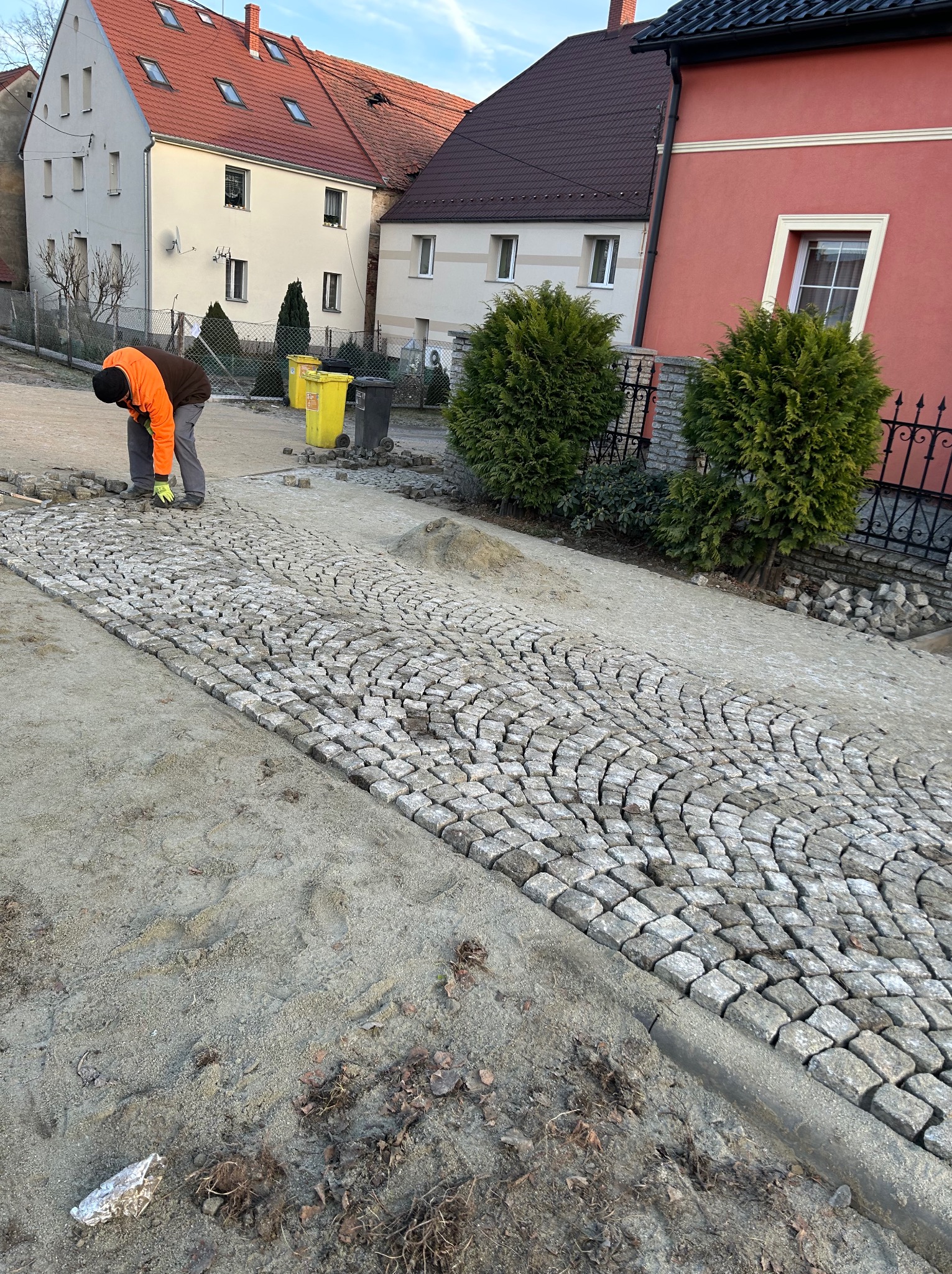 Układanie kostki granitowej na posesji. Widoczne ułożone fragmenty i pracownik w pomarańczowej kurtce. W tle domy i roślinność.