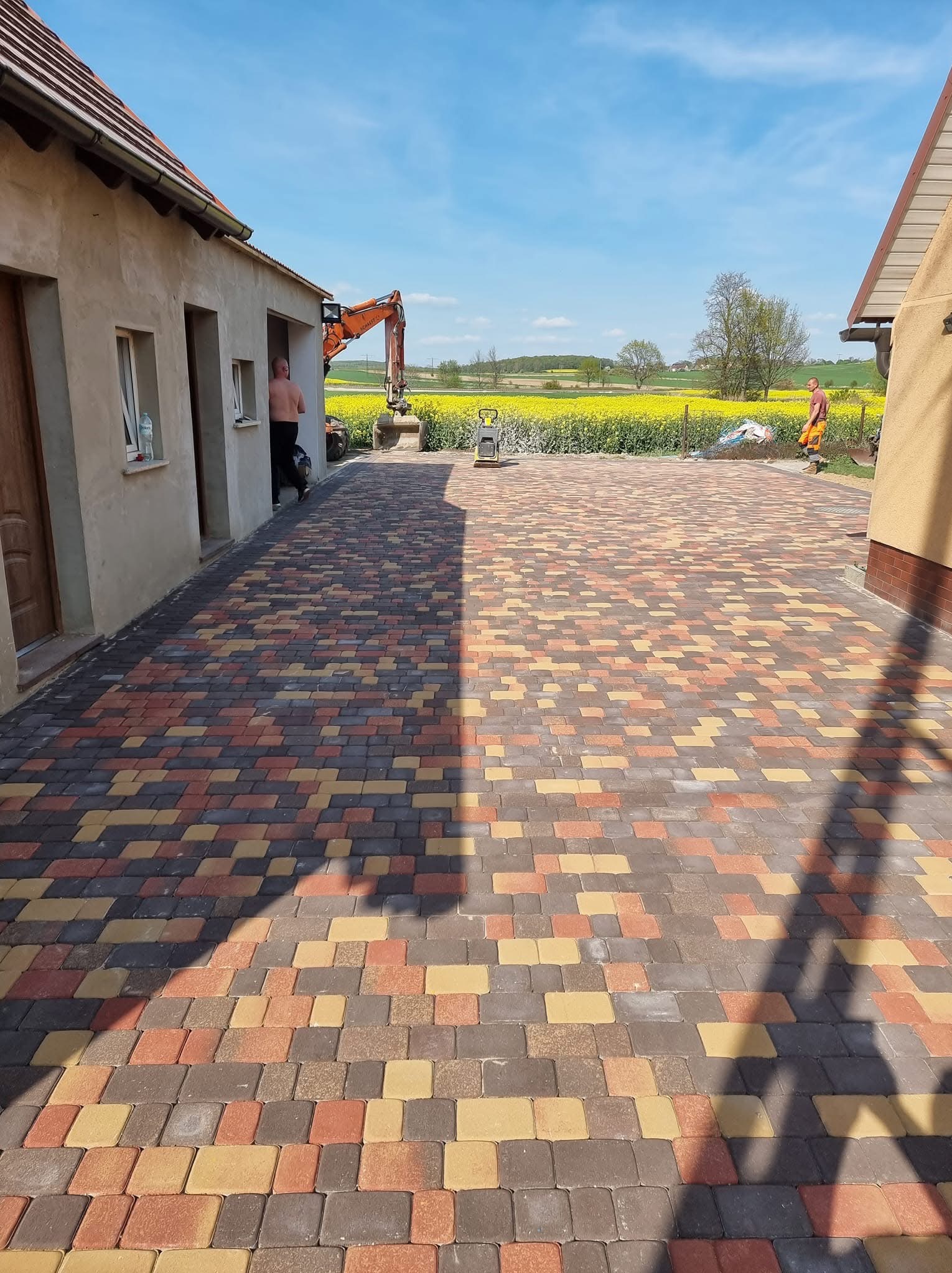 Kolorowa kostka brukowa tworzy wzór na podjeździe. W tle żółte pole, budynki i pracownicy. Widoczny sprzęt budowlany: koparka i zagęszczarka.