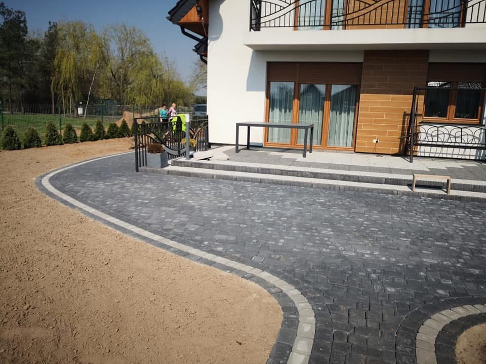 Ułożona kostka brukowa w kolorze grafitowym z jasną obwódką, prowadząca do domu z balkonem. Widoczny fragment trawnika i osoby w tle.