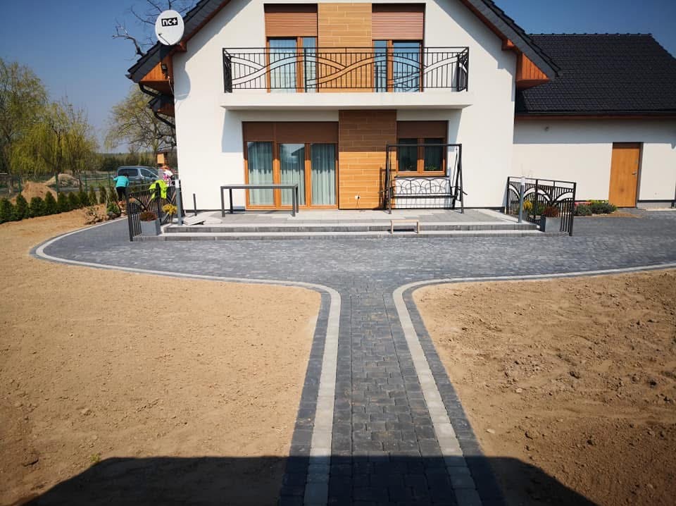 Nowoczesny podjazd z szarej kostki brukowej prowadzący do domu z balkonem. Widoczny fragment ogrodu z młodymi tujami i pracownicy w tle.