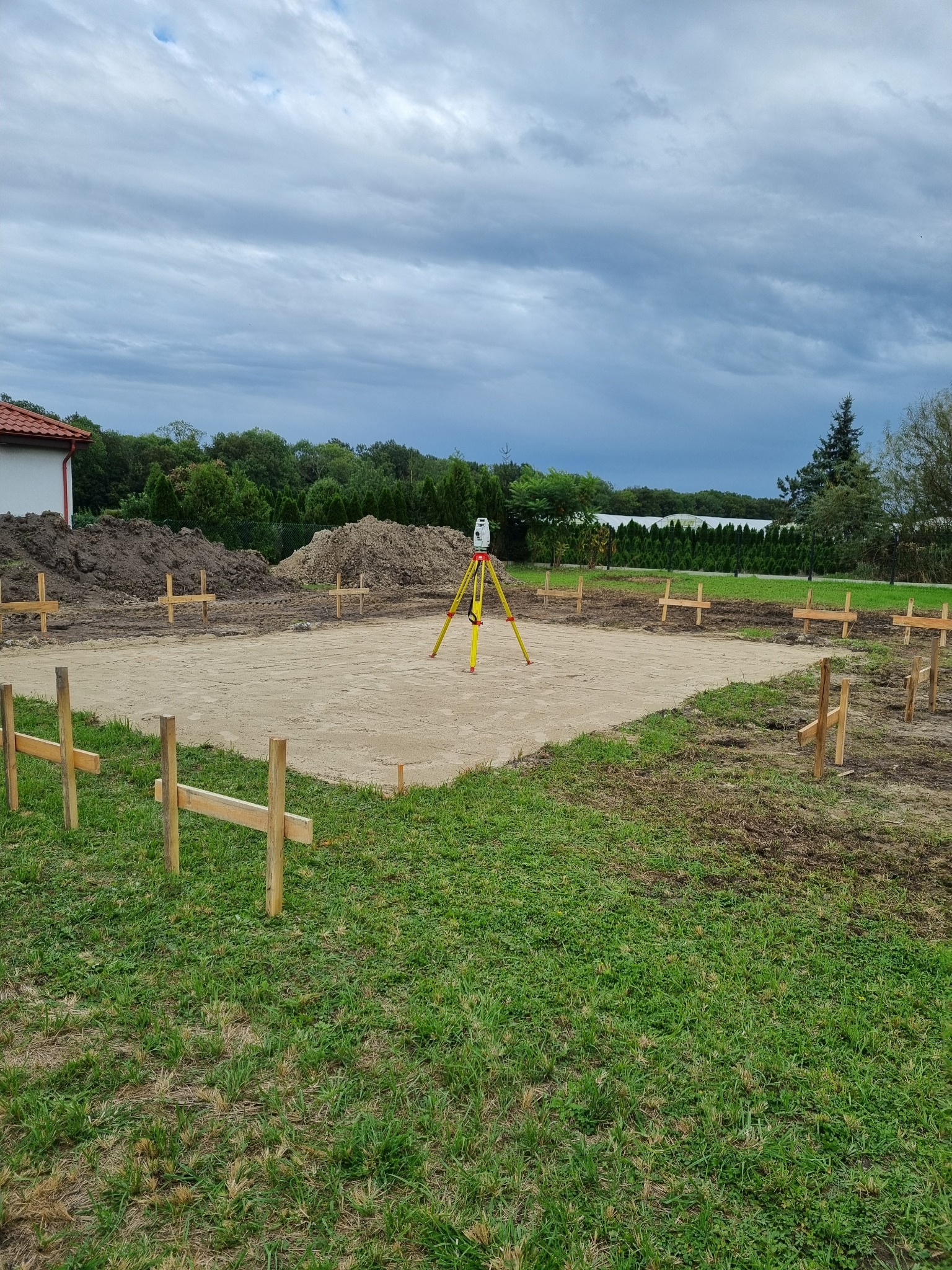 Wytyczanie fundamentów pod budowę domu. Niwelator geodezyjny na statywie, drewniane paliki wyznaczające obrys. Przygotowany teren pod wylanie fundamentów.