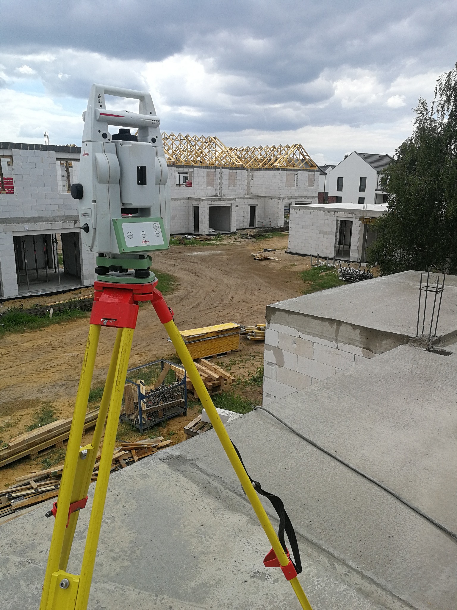 Tachimetr Leica na żółtym statywie na tle budowy osiedla domów jednorodzinnych w stanie surowym otwartym. Widoczne zbrojenia i więźby dachowe.