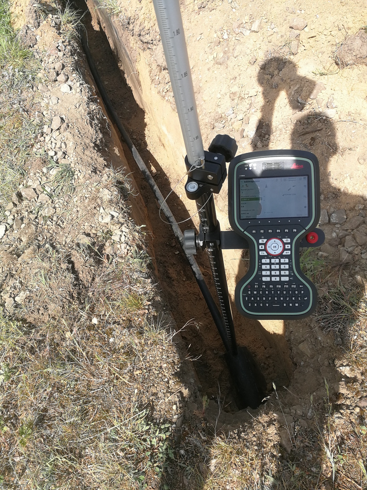 Pomiar wykopu pod instalację: niwelator z kontrolerem Leica, łata geodezyjna, cień osoby. Precyzyjne wyznaczenie trasy przebiegu instalacji w ziemi.