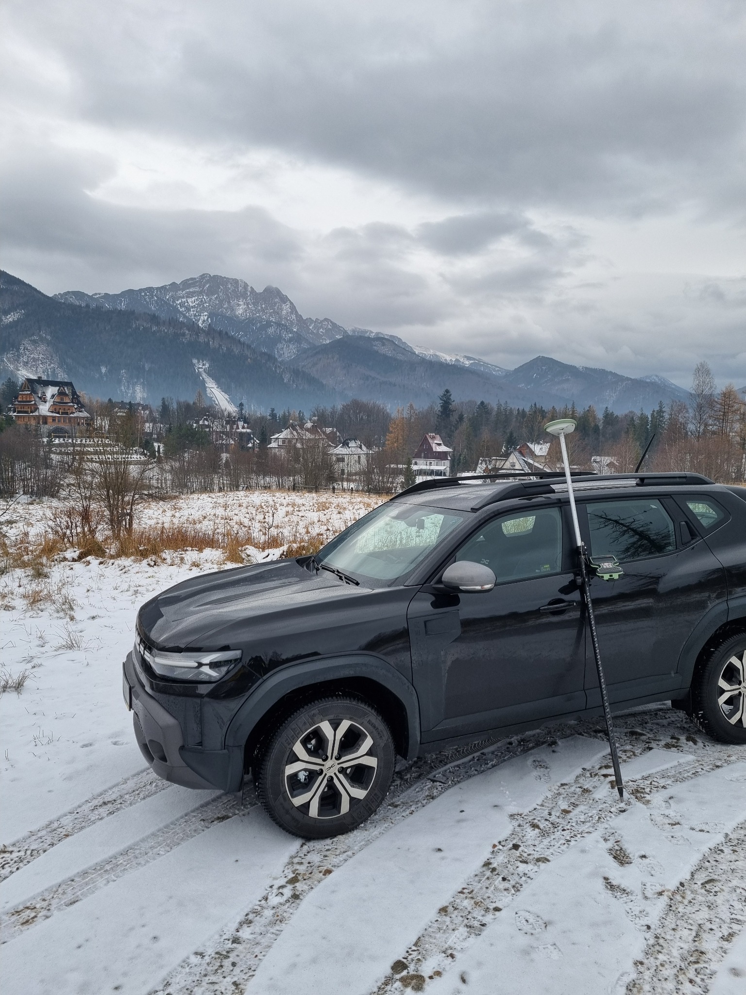Czarny SUV z anteną GPS na tle zimowego krajobrazu górskiego. Ślady opon w śniegu sugerują terenowe pomiary. Pochmurne niebo, zabudowania w oddali.