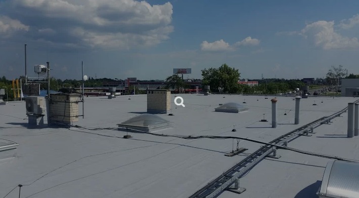 Szary, płaski dach budynku komercyjnego z widocznymi świetlikami, kominem, instalacjami i elementami wentylacji. W tle zabudowa miejska i błękitne niebo.