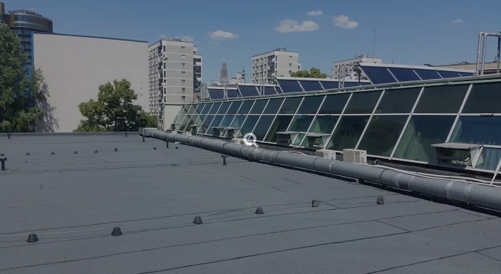 Płaski dach pokryty papą termozgrzewalną z widocznymi elementami wentylacji i klimatyzacji, na tle zabudowy miejskiej w słoneczny dzień.