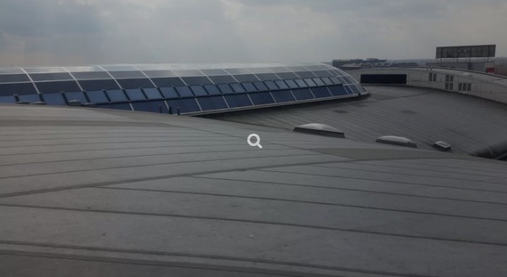 Szary, blaszany dach przemysłowy z świetlikami i przeszkloną kopułą w oddali, widok z góry przy pochmurnej pogodzie.