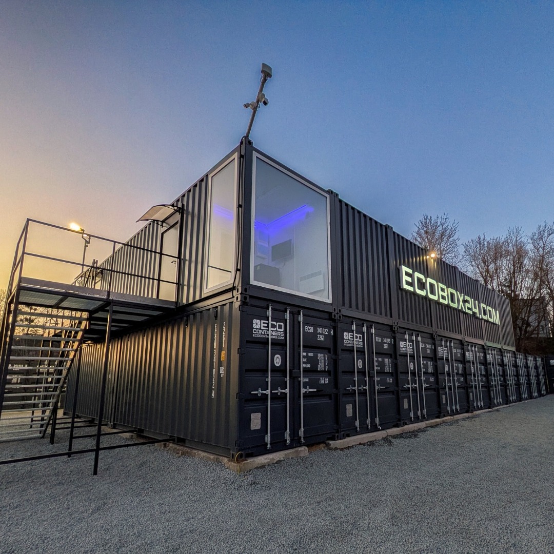 Nowoczesny magazyn kontenerowy ECOBOX24.com z oknem i schodami zewnętrznymi, oświetlony, na żwirowym podłożu, w tle drzewa i błękitne niebo o zmierzchu.