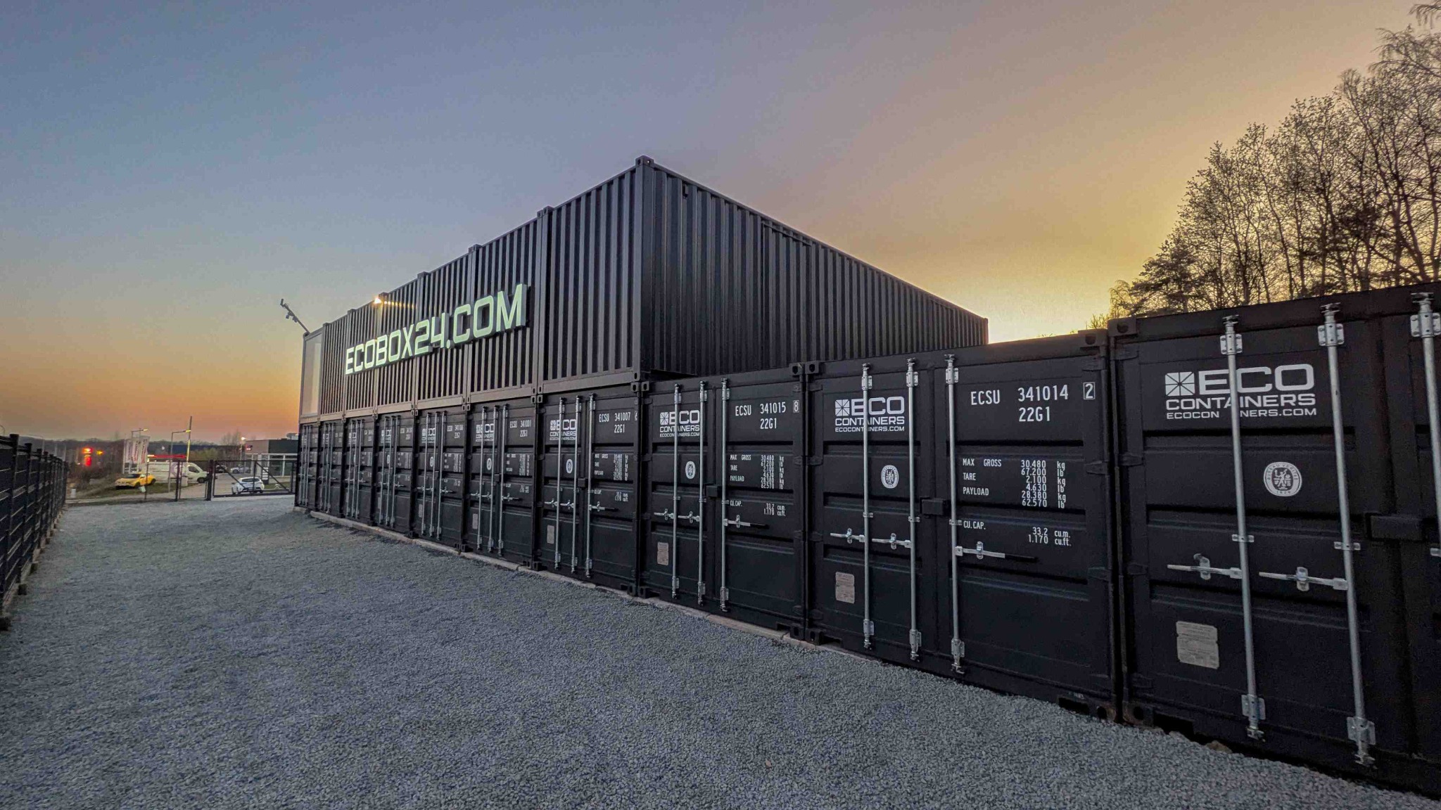 Czarne kontenery magazynowe ECOBOX24.COM ustawione jeden na drugim, widoczne napisy i logo firmy. Szary żwir na ziemi, wieczorny krajobraz w tle.