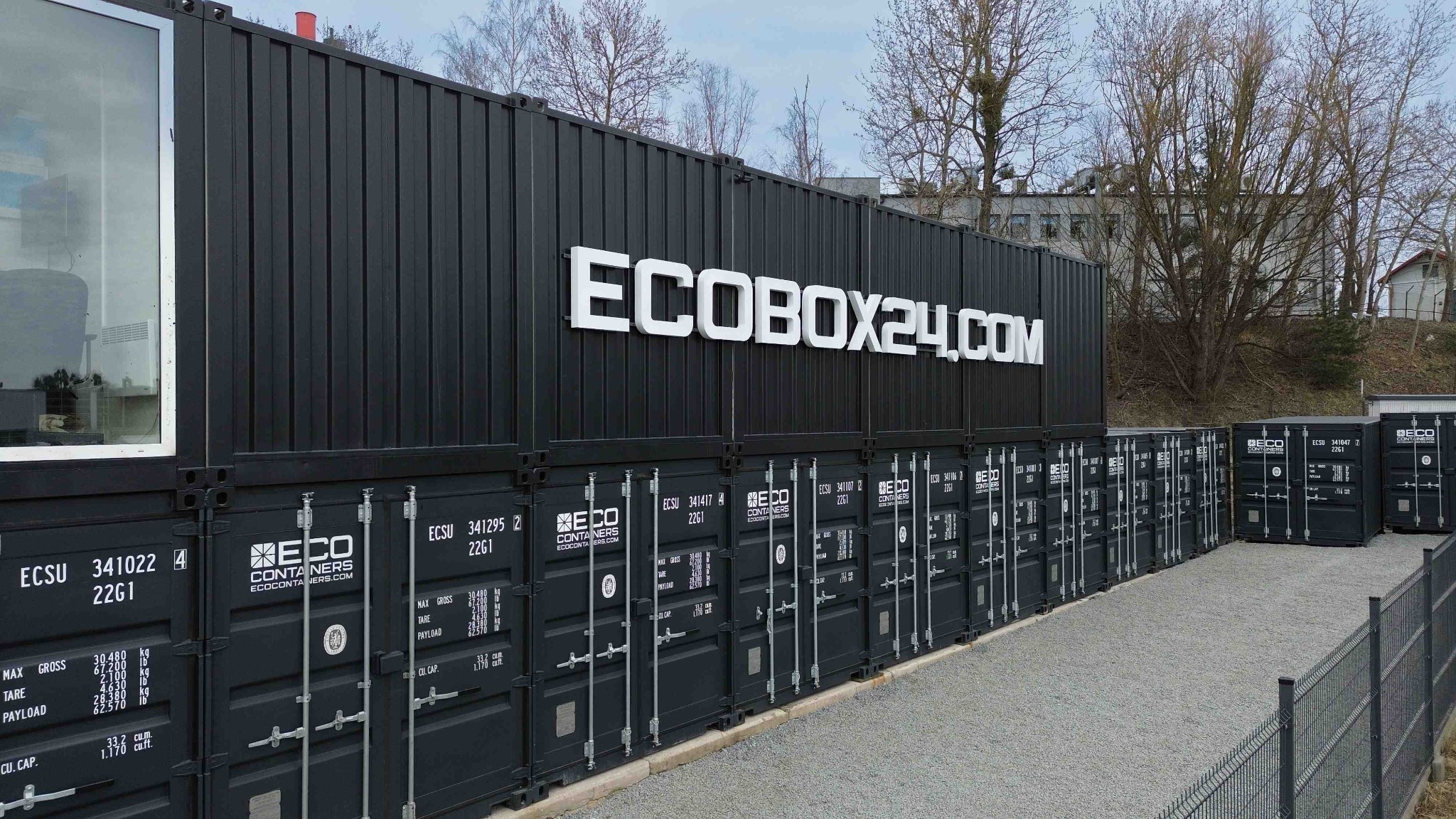 Czarne kontenery magazynowe ECOBOX24.COM ustawione jeden na drugim, z widocznym oknem w jednym z nich i szarym żwirem na ziemi.