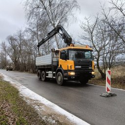 Wespol - Żółta ciężarówka z dźwigiem w Szadku na drodze, w tle drzewa bez liści i inny pojazd. Widoczny znak drogowy.