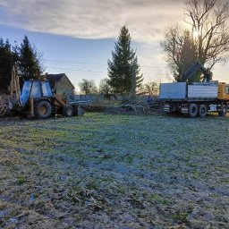 Wespol - Żółty traktor z łyżką i ciężarówka z dźwigiem na polu po wycince drzew. Słońce przebija przez korony drzew. Prace ziemne w toku.