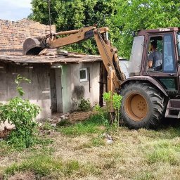 Wespol - Żółto-brązowa koparko-ładowarka CASE podczas rozbiórki małego, murowanego budynku z ceglanym fragmentem ściany. Operator w kabinie. Widoczne elementy roślinności.