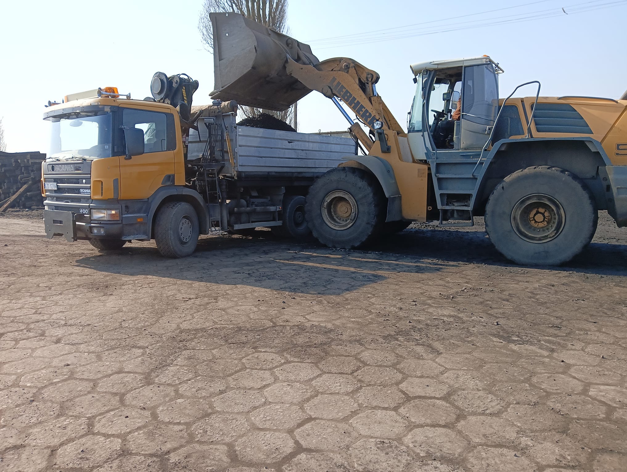 Żółta ładowarka Liebherr nabiera ziemię i wsypuje ją do skrzyni ciężarówki Scania na placu budowy. Widoczne opony i kabina operatora.