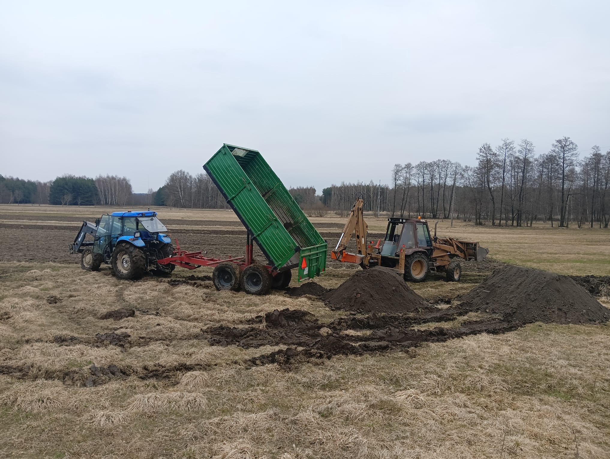 Niebieski traktor z zieloną przyczepą wywrotką i koparko-ładowarka na polu z pryzmami ziemi. Prace ziemne w toku, widok na pole i drzewa w tle.