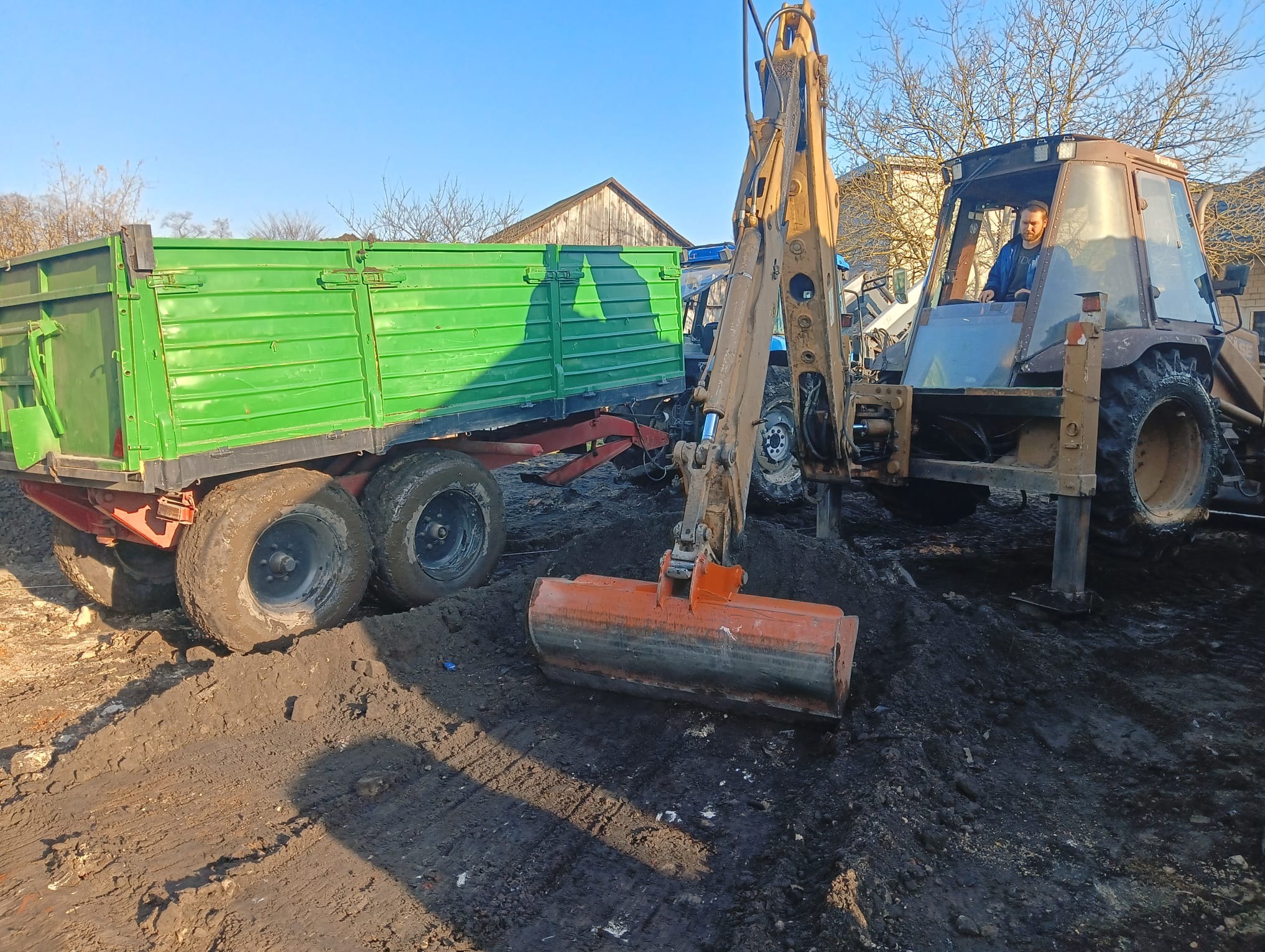 Praca ziemna: Traktor z łyżką kopiącą obok zielonej przyczepy na błotnistym terenie w słoneczny dzień. Operator w kabinie.