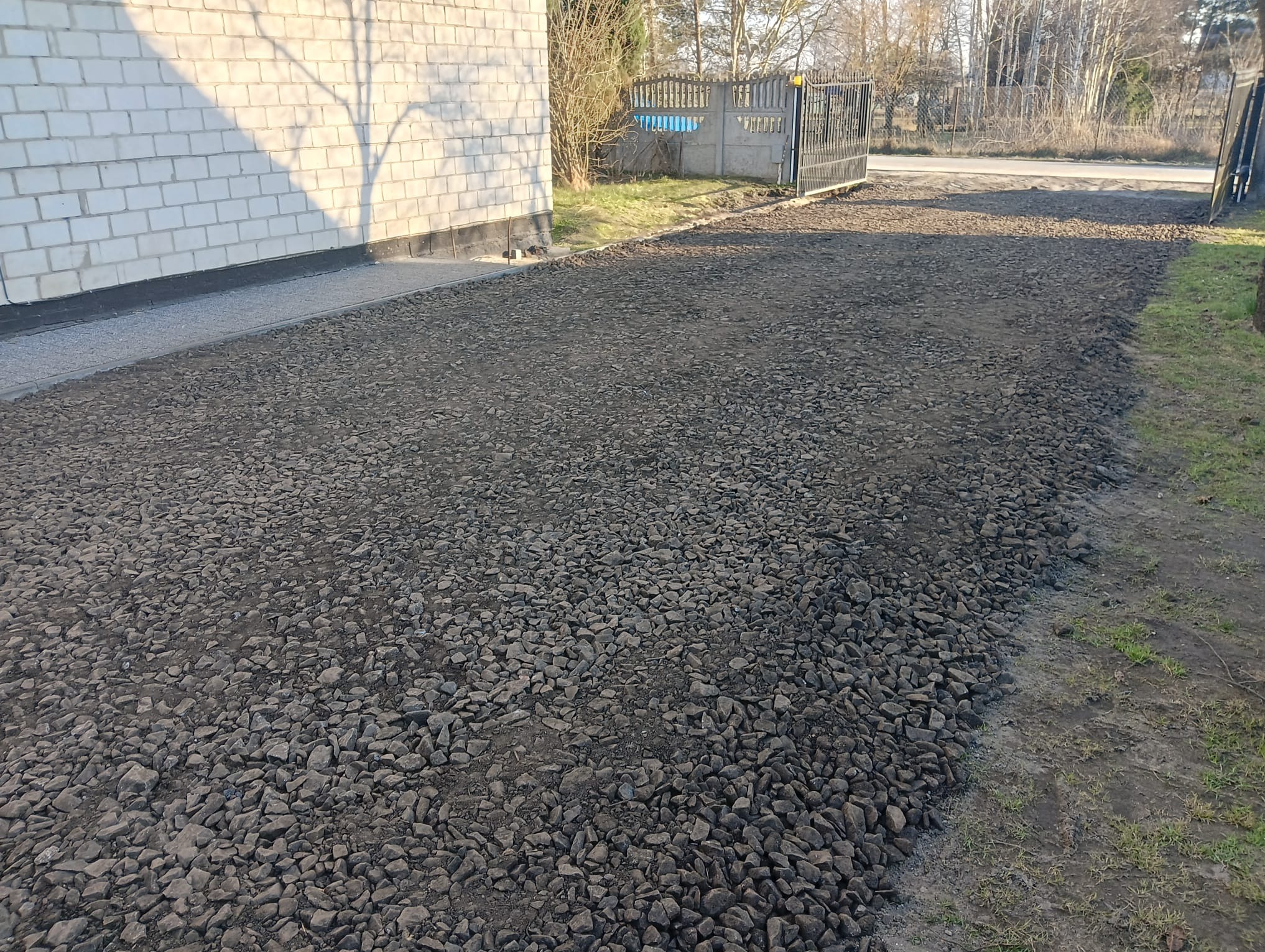 Utwardzona droga do posesji z bramą, widok z bliska na szarą nawierzchnię z kamieni. W tle budynek z białej cegły i drzewa.
