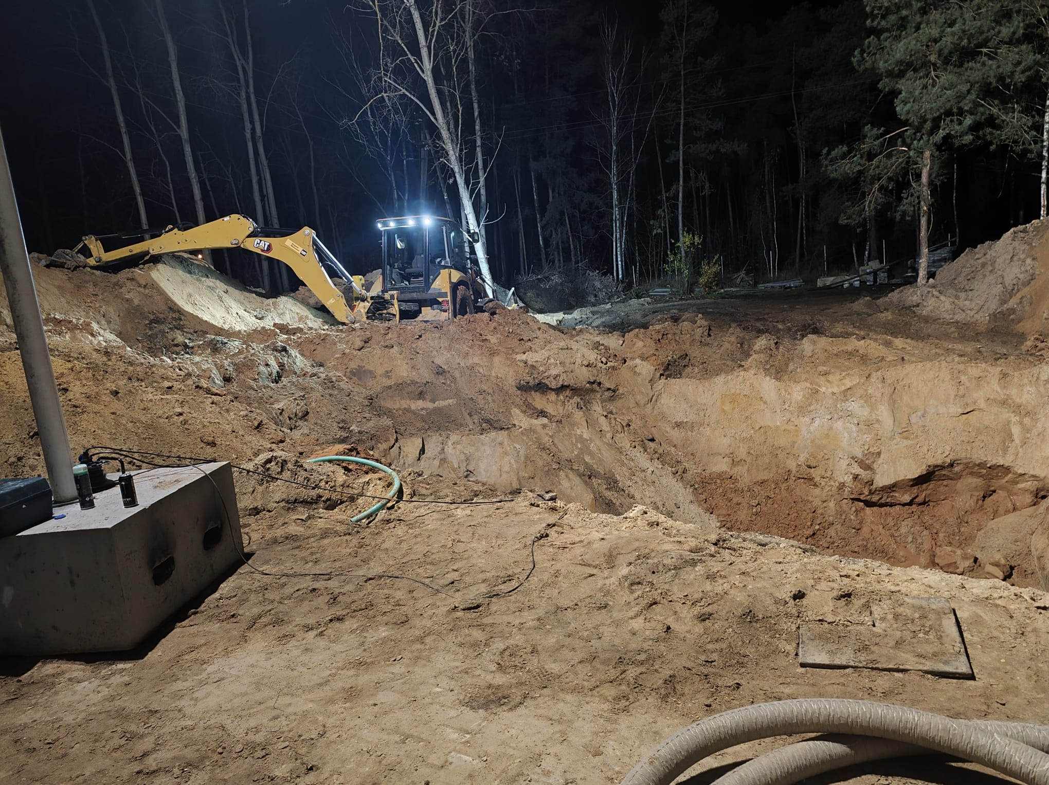 Żółta koparka CAT oświetla wykopany dół w nocy. Widoczne drzewa w tle i betonowy element na pierwszym planie. Prace ziemne przy sztucznym oświetleniu.